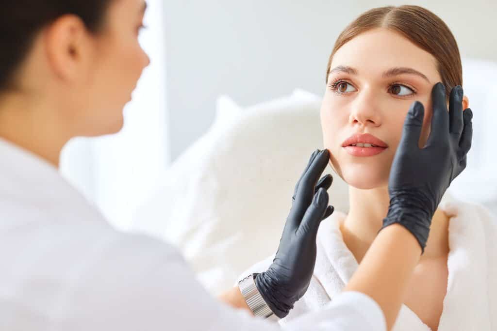 Cosmetologist examining face skin of young female client before proceeding to botox and laser treatment